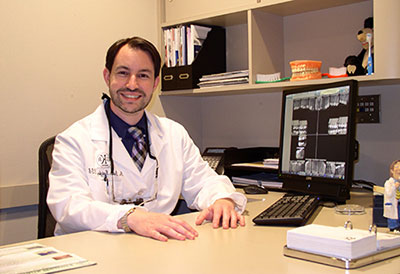 The image features a man sitting at a desk in an office setting, wearing a white lab coat and smiling.