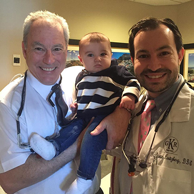 The image shows a man holding a baby, with another man in the background wearing a stethoscope and a name tag, suggesting they are in a medical setting.