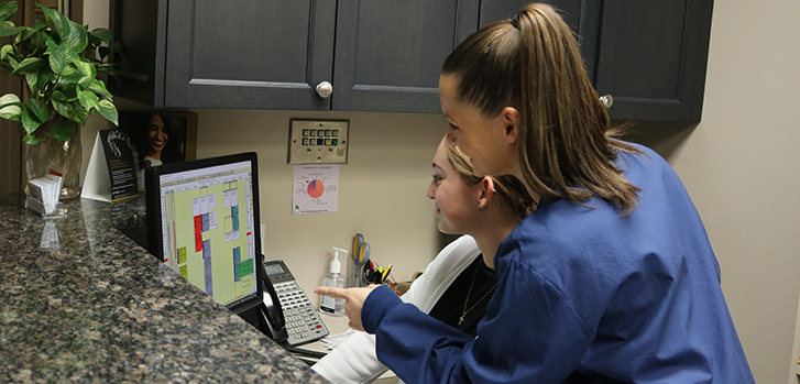 The image depicts a scene at a desk with a laptop, where an individual appears to be using the computer while another person is standing nearby, possibly engaged in conversation or observing the screen.