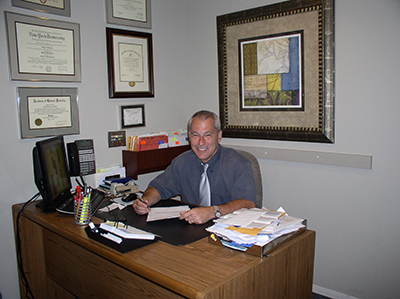 The image shows a man sitting at an office desk, smiling and signing papers with a pen in his hand.