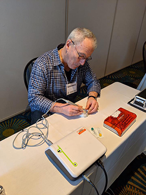 A man is seated at a table, working on a device with various electronic components and cables.