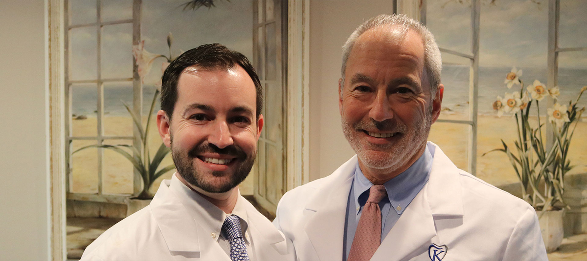 Two men standing side by side, one wearing a white lab coat and the other in a suit, both smiling at the camera.