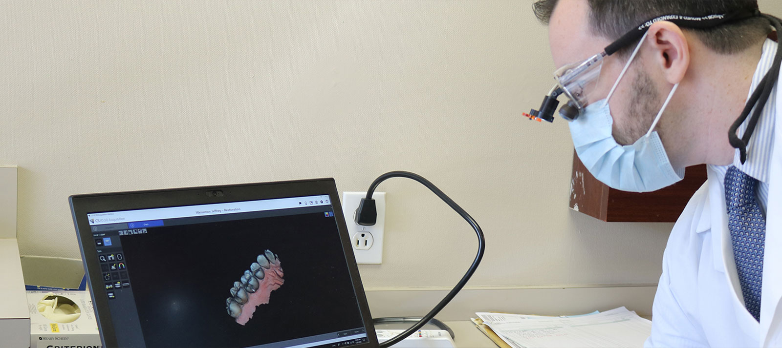 The image shows a person wearing a white lab coat and a face mask, sitting at a desk with a laptop screen displaying what appears to be a 3D model of an object. The individual is using a digital stylus to interact with the model on the screen.