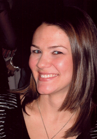 The image is a photograph of a smiling woman with short brown hair, wearing a black top and a necklace. She has a fair complexion and is looking directly at the camera.