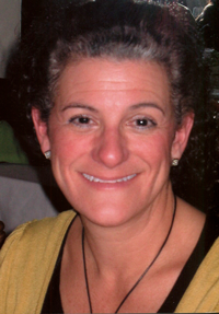 A woman with short gray hair, wearing a yellow top and a necklace, smiling at the camera.