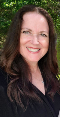 The image shows a woman with long hair, smiling at the camera. She has dark hair and is wearing makeup. The background is out of focus but appears to be an outdoor setting with natural light.