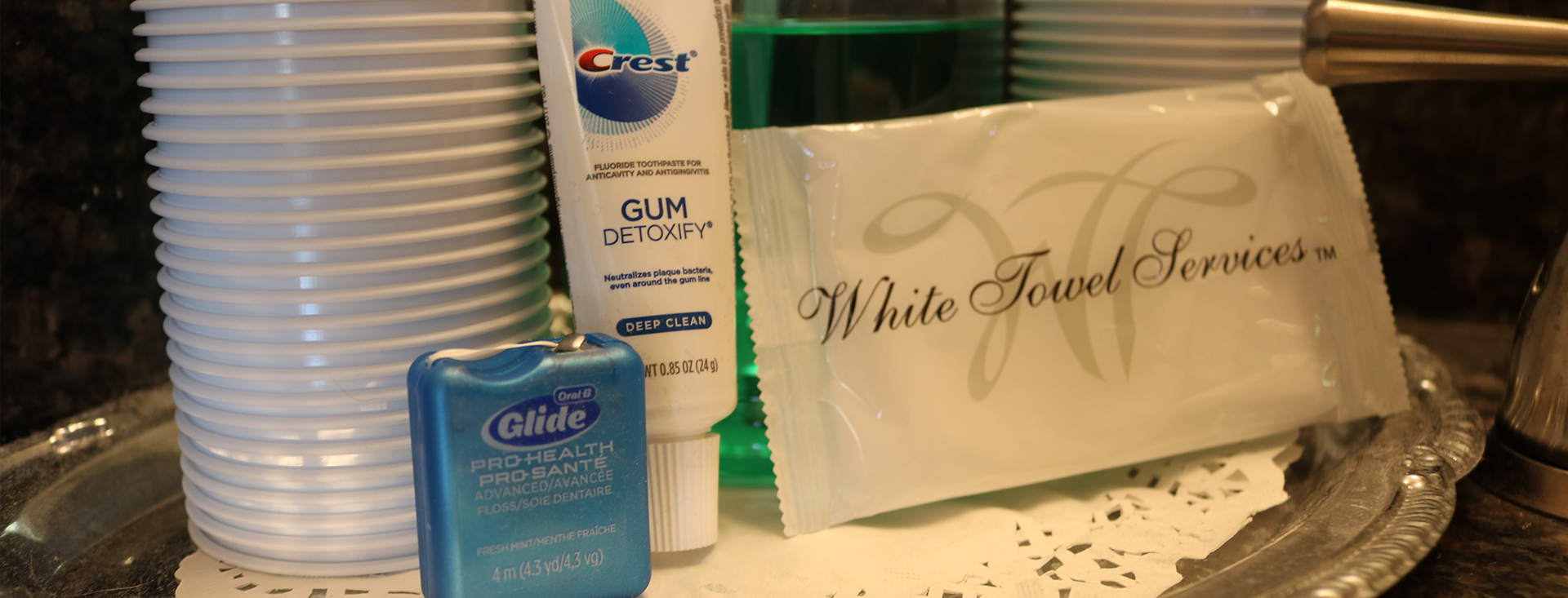 The image displays a collection of personal care items and cleaning supplies, including toothbrushes, toothpaste, mouthwash, and other hygiene products, arranged on a tray with a sign that reads  White Glove Services.