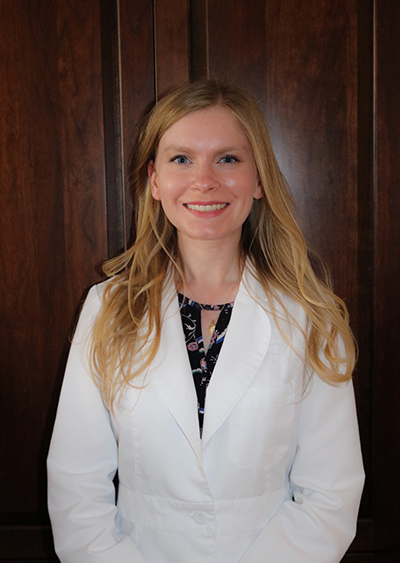 The image shows a woman standing in front of a paneled wall, wearing a white lab coat and smiling at the camera.