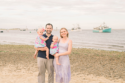 A family of four on a beach, with a man, woman, baby, and toddler posing for a photo.