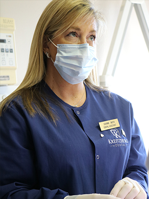 A woman in a blue scrub uniform, wearing a surgical mask and gloves, is seated at a desk with various medical equipment.