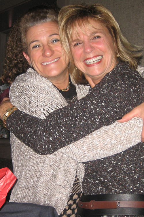 The image shows two women embracing each other in a warm hug, with one woman wearing a festive red and white scarf.