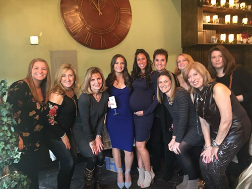 The image shows a group of people posing together at an event, with a pregnant woman in the center and another woman to her right. They are gathered around a table with a bottle on it, and there is a clock visible in the background.