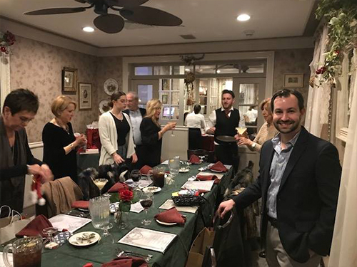 The image shows a man standing at the end of a dining table in what appears to be a festive setting, possibly a holiday gathering or celebration, with people seated around the table engaged in conversation.