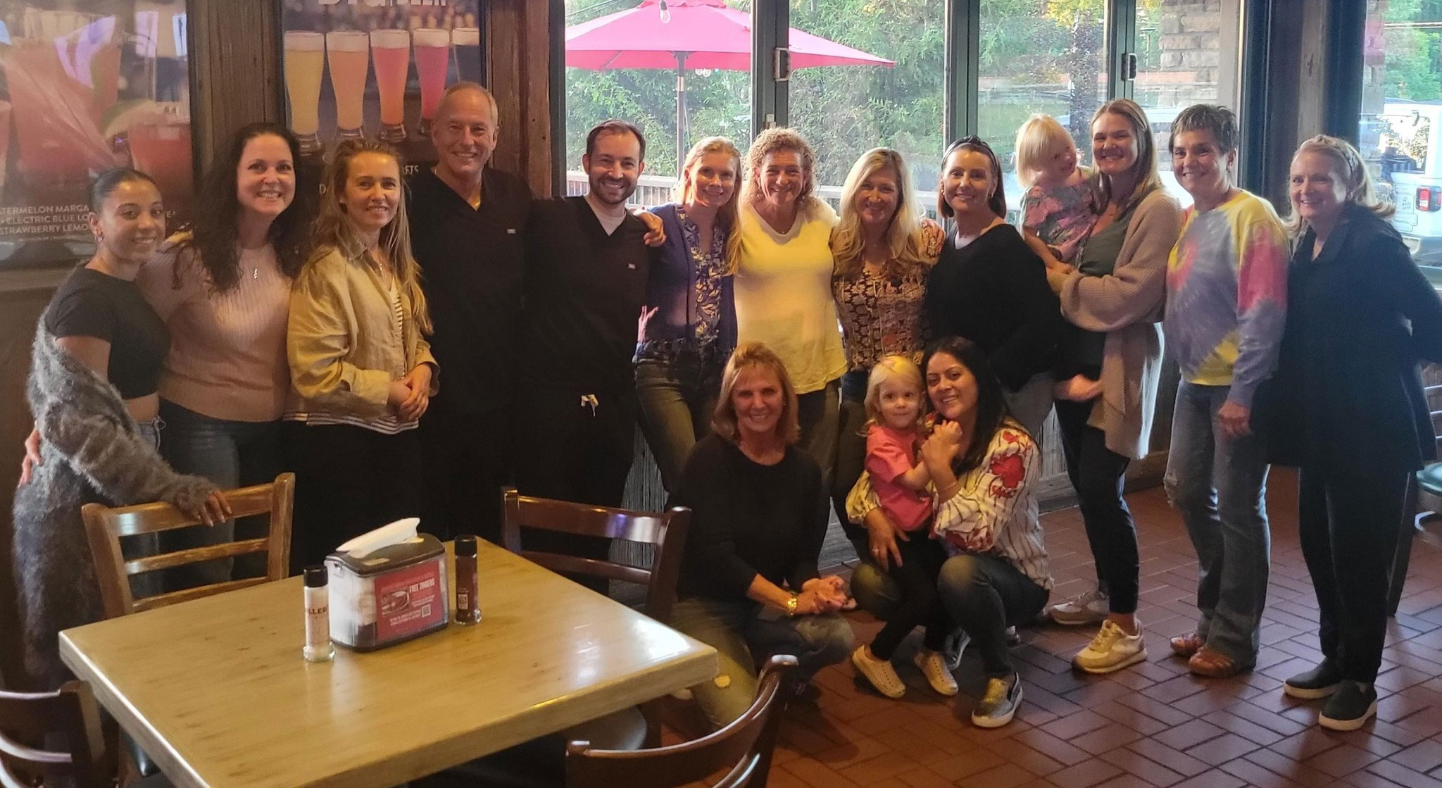 A group of people posing for a photo in a restaurant setting, with individuals seated around dining tables and standing behind them.