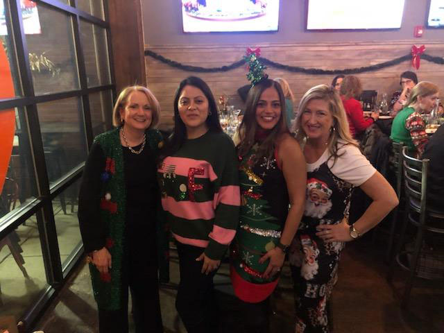 The image shows three women posing together at a festive event, with one woman in the center wearing a holiday-themed sweater and two others on either side dressed in matching outfits.