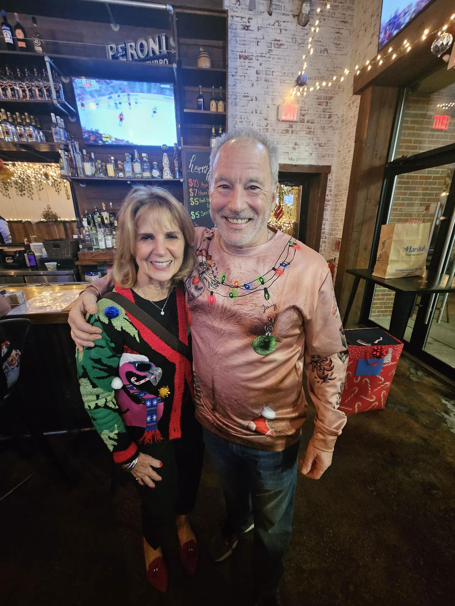 A man and a woman, both wearing festive holiday outfits, posing together for a photograph at what appears to be a bar or restaurant.