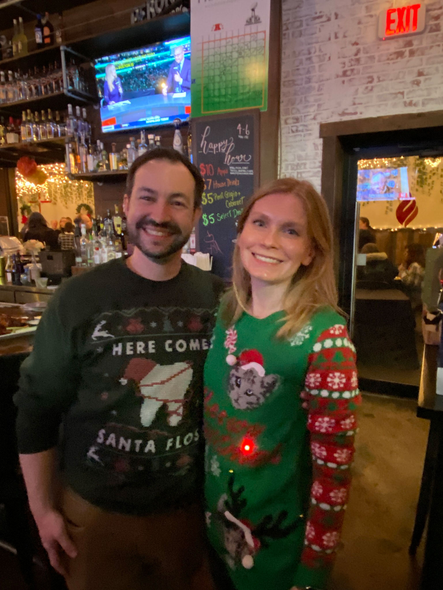 The image shows a man and woman posing for a photo at what appears to be a bar or restaurant. They are both smiling and wearing festive holiday attire, with the man in a sweater that says 'Here Comes Santa' and the woman in a Santa Claus-themed outfit.