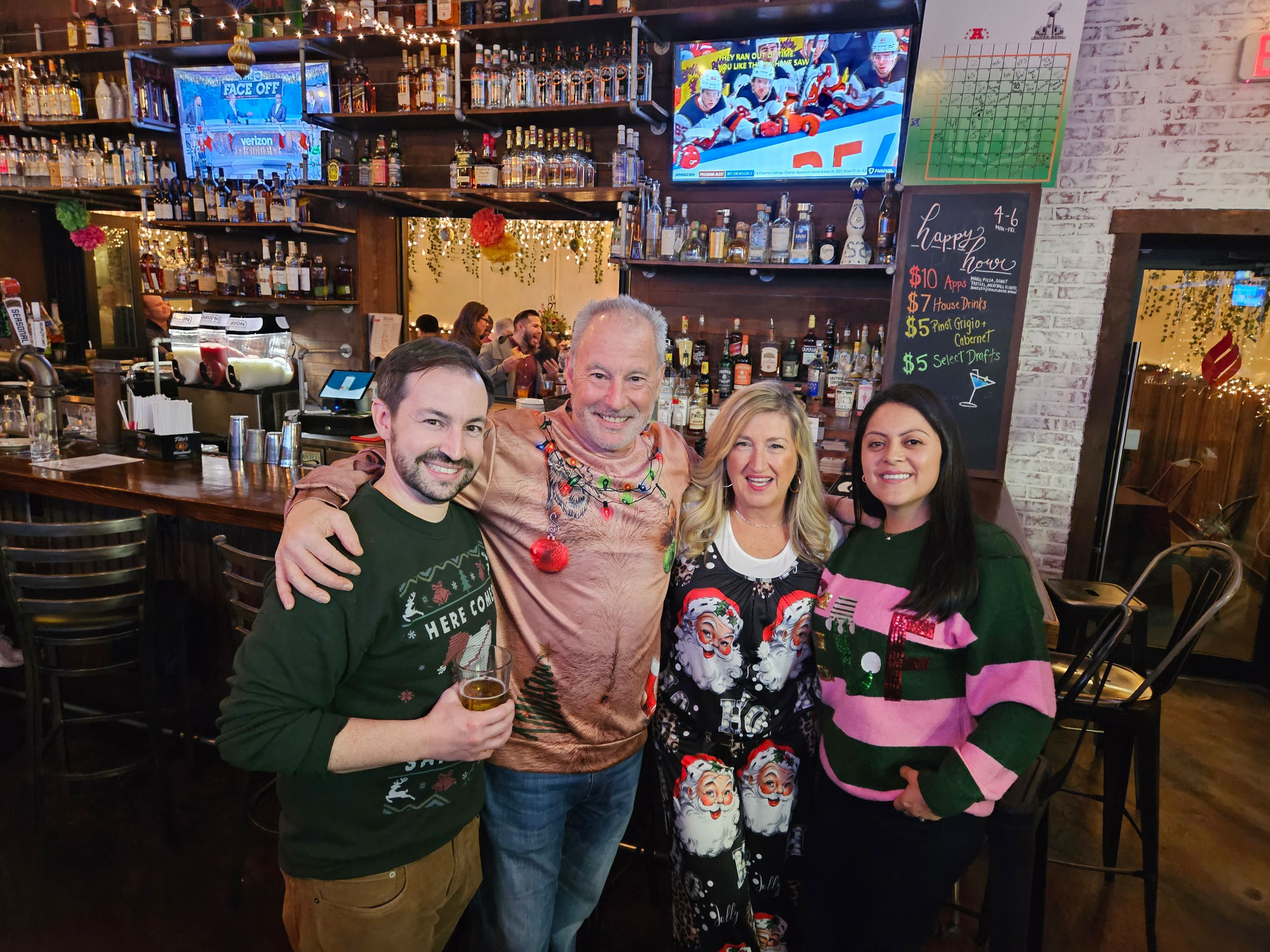 The image shows a group of people, likely at a social gathering or event, posing for a photo with a festive backdrop that includes Christmas decorations and a sports-themed sign.