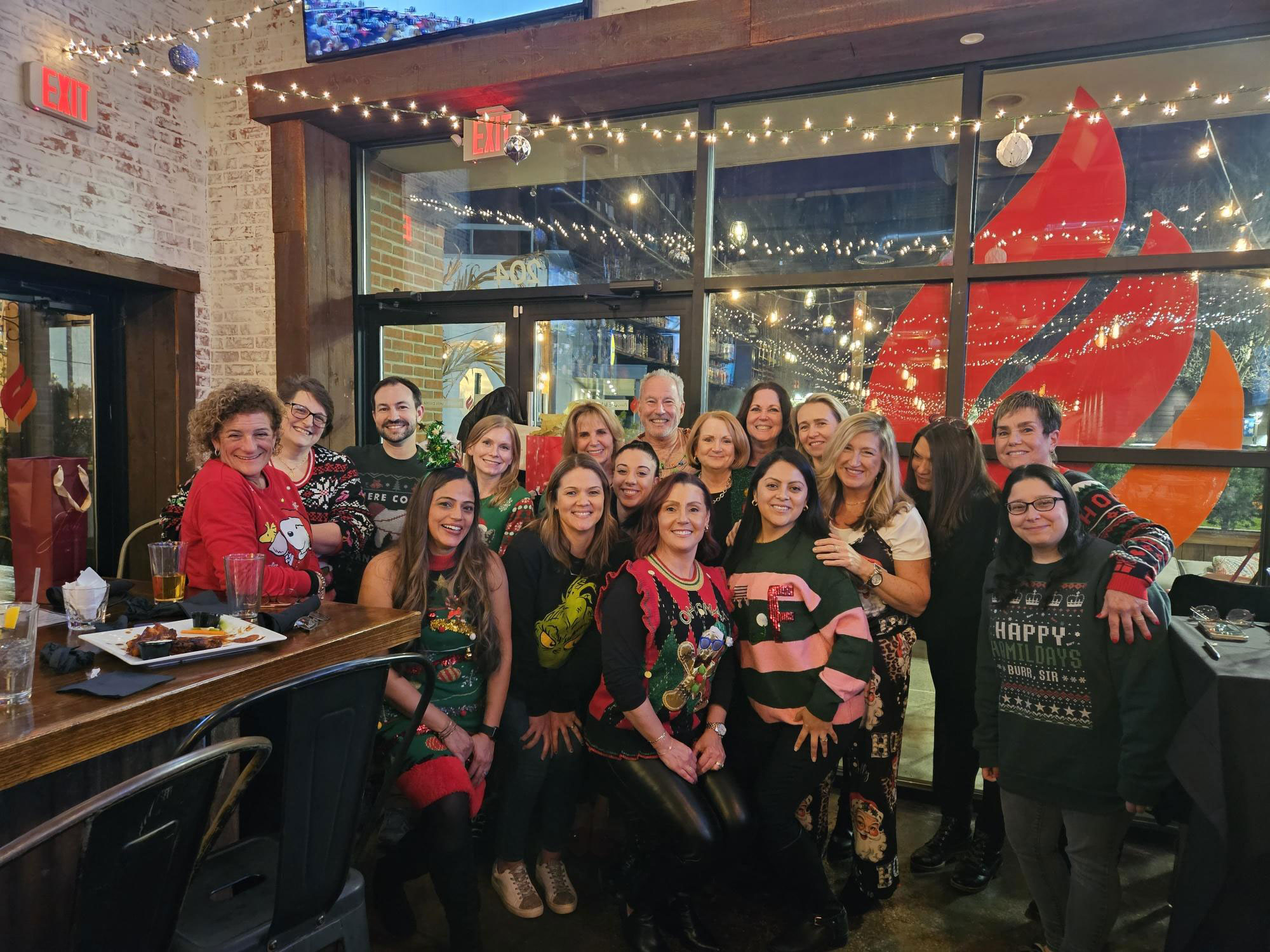 The image is a group photo of a festive gathering, likely a holiday party or celebration, with people dressed in seasonal attire and posing together in front of a restaurant interior.