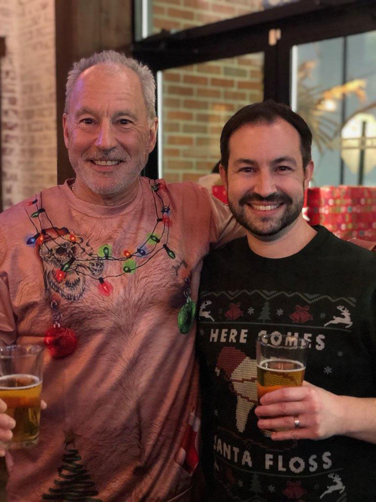 In the image, two men are standing together, one wearing a festive holiday sweater and the other in a Santa Claus onesie. They are holding drinks, smiling, and posing for the camera.