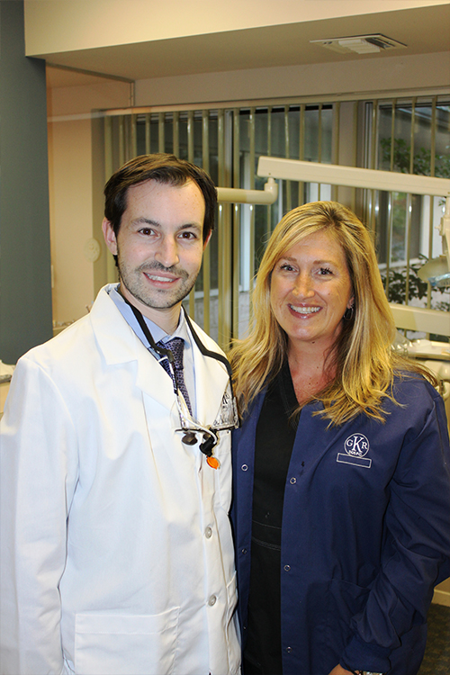 The image shows a man and a woman posing together. The man is wearing a white lab coat, suggesting he may be a scientist or medical professional, while the woman appears to be in casual attire. They are standing indoors with a neutral background that does not provide any specific context about their location.