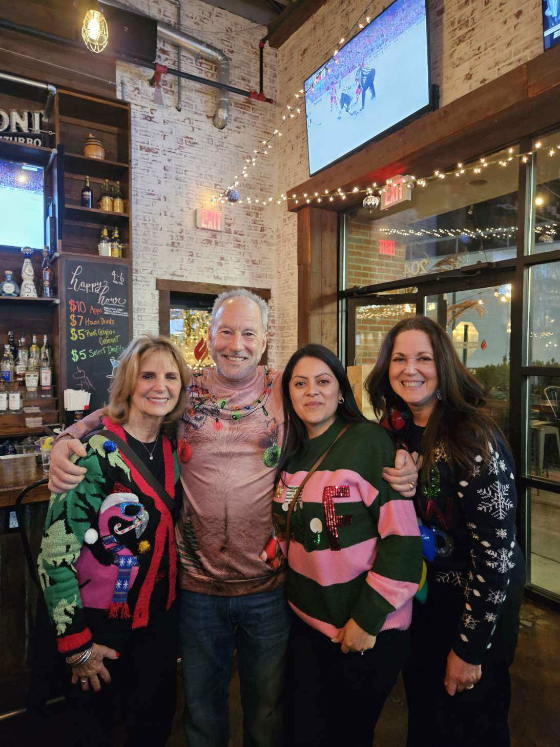 The image shows four people posing together in a festive setting, likely a holiday party or gathering. They are dressed in seasonal attire and stand in front of a decorated background with Christmas lights.