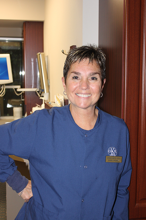 The image shows a woman wearing a blue scrub top, standing in an indoor setting that appears to be a medical or dental office, smiling and looking directly at the camera.