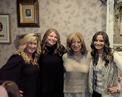 The image depicts four women posing together for a photograph, with one woman standing slightly apart from the group. They are all smiling and appear to be in a social setting, possibly a party or gathering.