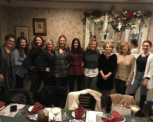 A group of women posing for a photo at a formal event, standing in front of a dining table with chairs and a festive atmosphere.