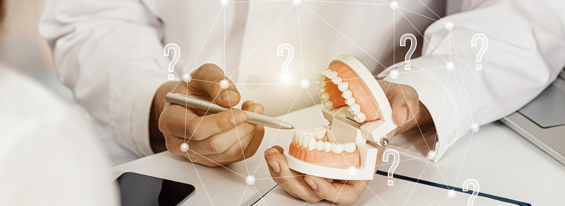 The image shows a person s hand holding a model of a human mouth with teeth, while another person is seated at a desk with a laptop and various papers.