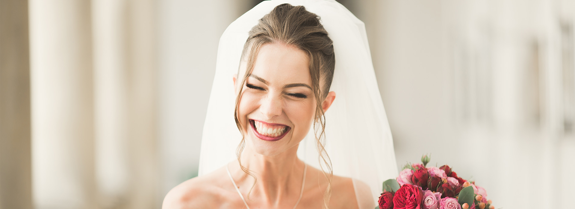 A bride wearing a white veil and holding a bouquet, smiling at the camera.