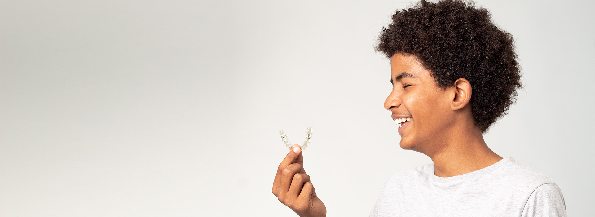 The image shows a person holding an object, smiling and looking at the camera. They are indoors with a plain background.