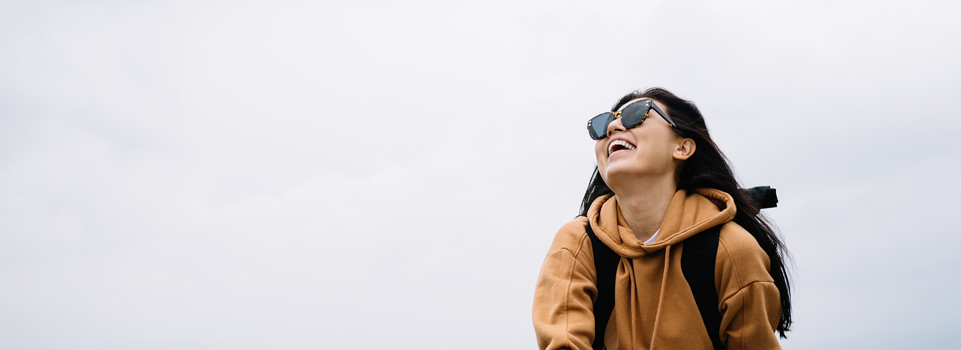 A person in a hoodie, sunglasses and backpack, smiling against a cloudy sky.