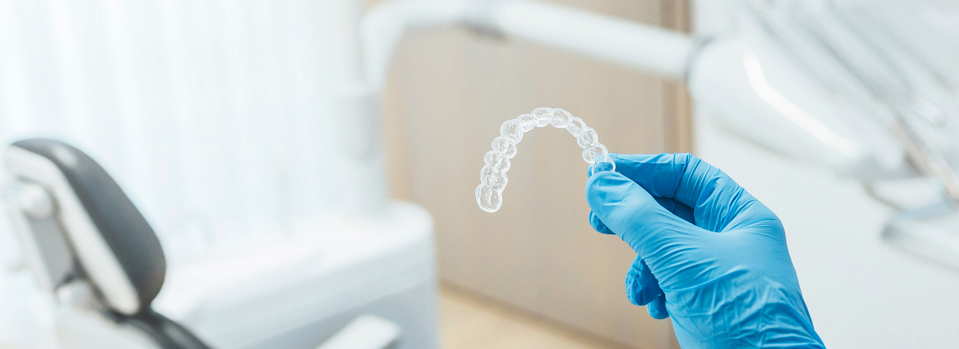 A close-up of a transparent dental impression material being held by a hand wearing blue gloves in front of a white dental chair and equipment.