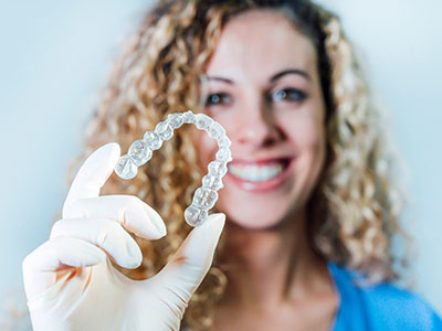 A smiling woman wearing a white glove holds up a transparent retainer with her left hand.