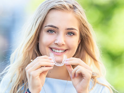 A young woman with blonde hair is smiling and holding a transparent dental retainer or aligner with her hand.