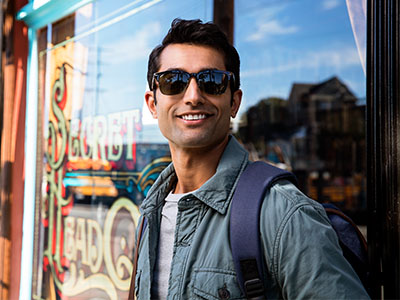 A man wearing sunglasses and a backpack, standing in front of a store window with a sign that reads  Sunday .