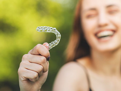 The image shows a young woman holding up a clear orthodontic aligner with her left hand, smiling and looking towards the camera.