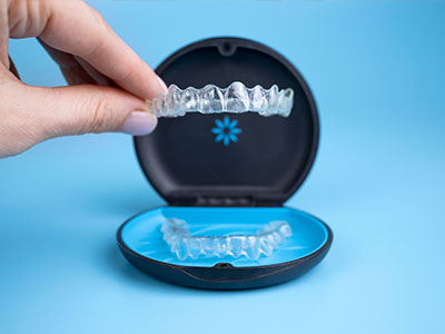 The image shows a hand holding an open dental retainer case with a blue case lid, revealing transparent aligners inside.