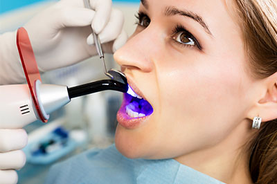 The image shows a person sitting in a dental chair with their mouth open, receiving dental care. A dental hygienist is using an ultrasonic cleaning device to clean the teeth.