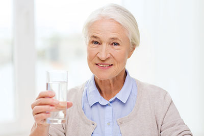 An elderly woman is holding a glass of water, smiling, and appears to be in a good mood.