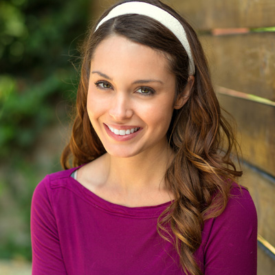 A woman with long brown hair is smiling at the camera, wearing a purple top and a headband.