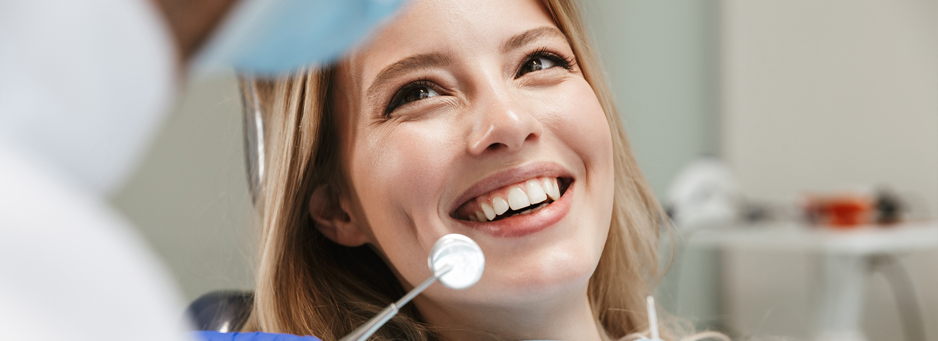 The image shows a woman smiling at the camera, with a dental professional in the background observing her smile.