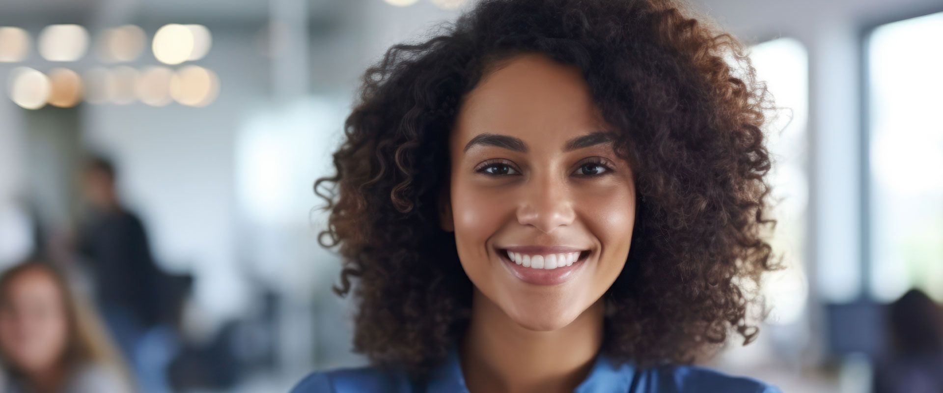 The image is a photograph of a smiling woman with curly hair, wearing a blue top, standing in an indoor office environment.