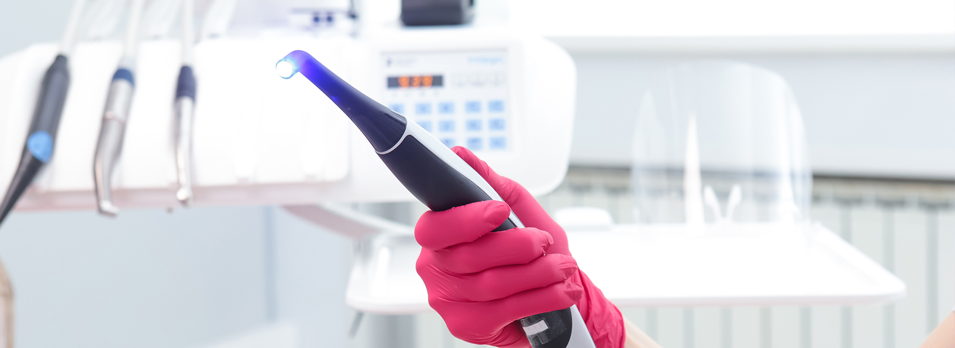 The image shows a person wearing pink gloves and holding a blue object, which appears to be an electronic device or gadget, while standing in what seems to be a laboratory setting with various equipment in the background.