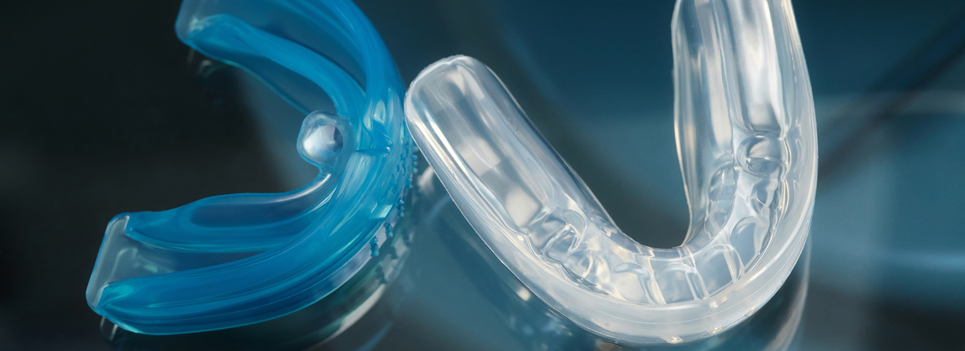 The image shows a close-up of two clear plastic dental retainers with blue accents, set against a blurred background that appears to be a dark surface.