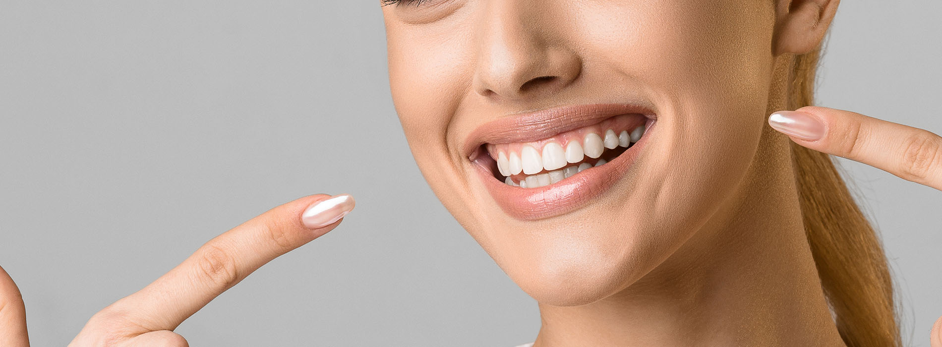 The image shows a woman with a radiant smile, holding her hands up to her cheeks in a gesture of surprise or delight. She has light skin and is wearing makeup, including lipstick. Her hair is styled in an updo, and she appears to be indoors with a neutral background.