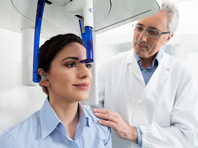 The image depicts a medical setting where a woman is seated in a dental chair, and a dentist is standing beside her, examining her teeth with the aid of a digital device.