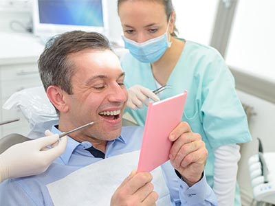 The image shows a man sitting in a dental chair, holding up a pink card or paper with a smile, while a female dental professional is seated behind him, examining his mouth.