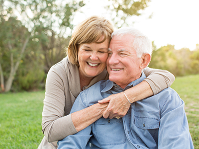 An elderly couple embracing in a park setting, with the man wearing a blue shirt and the woman in a white top.